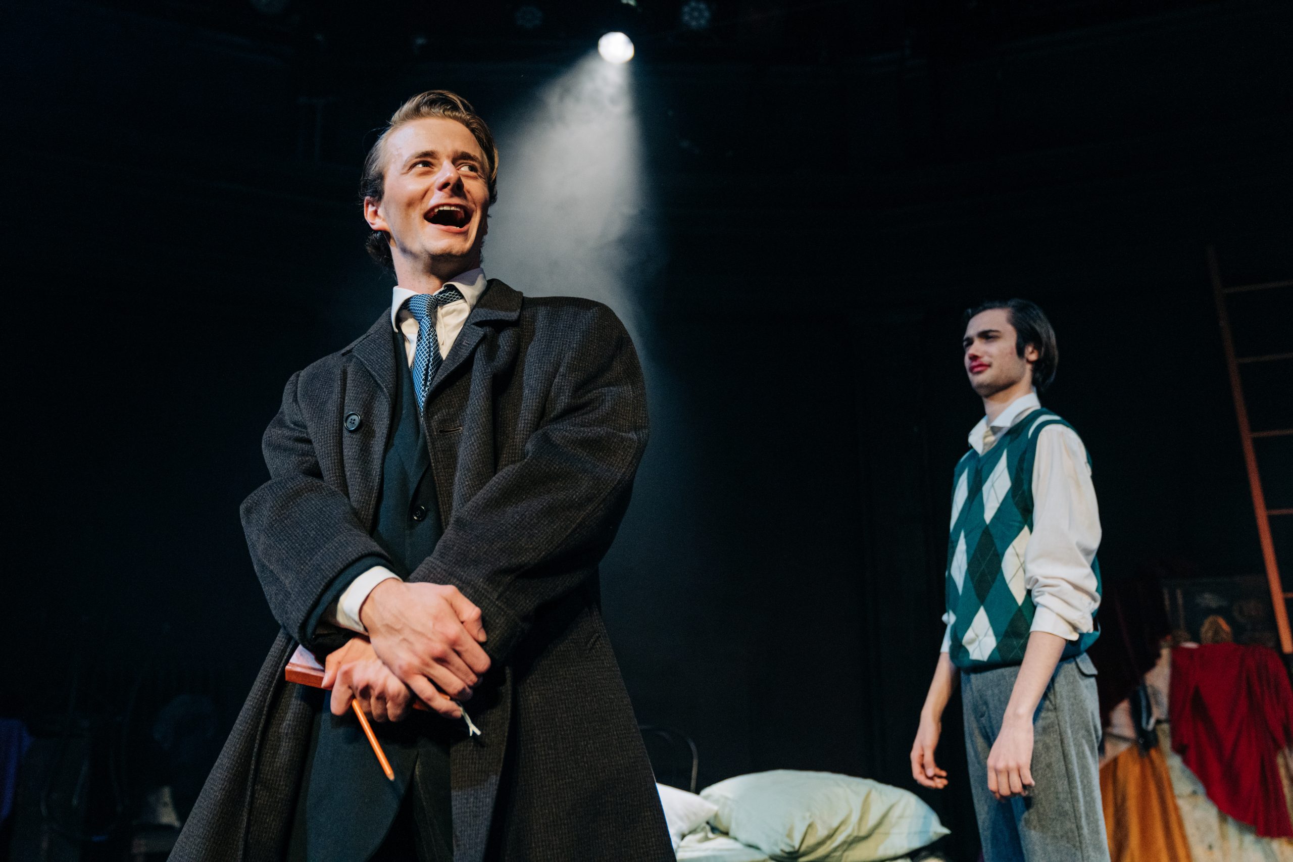 A young man in a trench coat stands center stage with a spotlight beaming down on him; he looks gleeful. A man being him looks very confused.