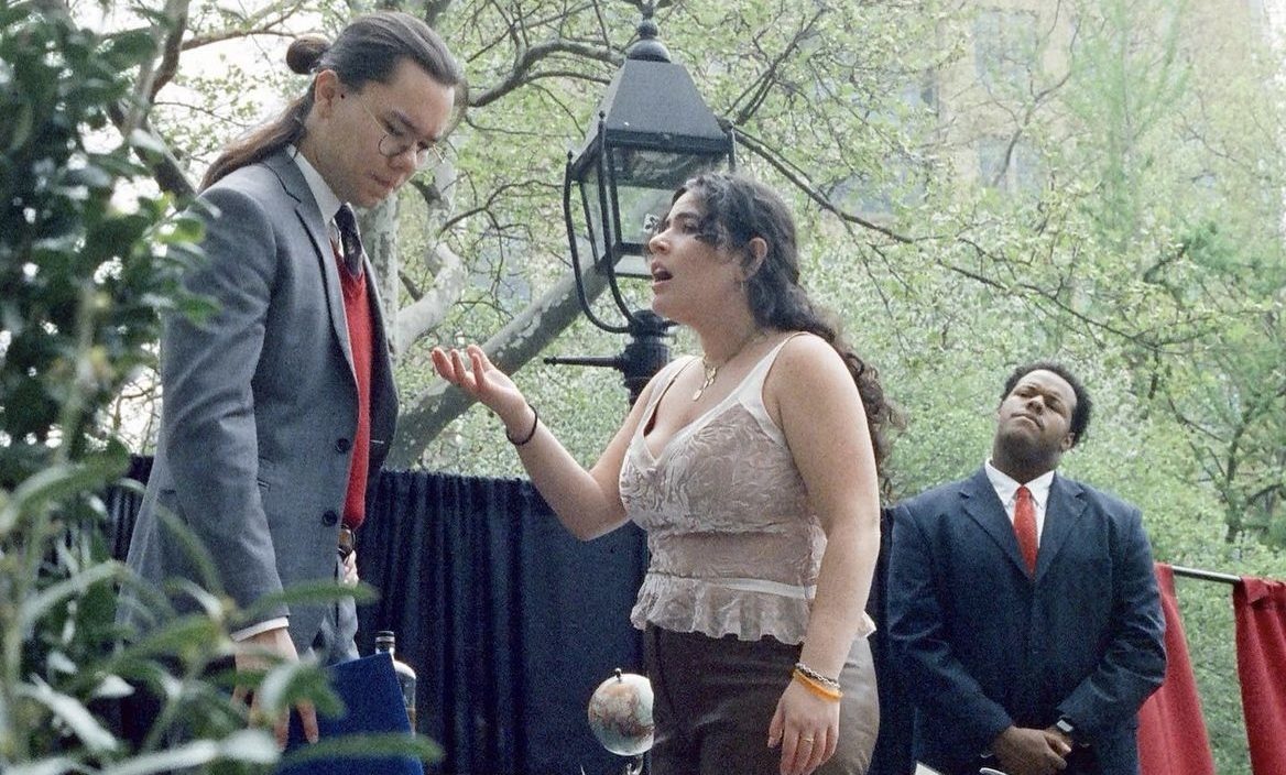 At an outdoor theater performance, a young woman argues with a disappointed man. Behind them, a young man watches with a thoughtful expression.