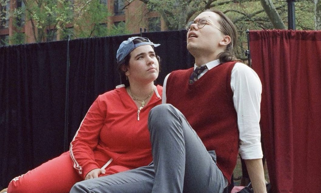 A young woman in a red tracksuit looks at a man with concern. He has a pained look on his face.