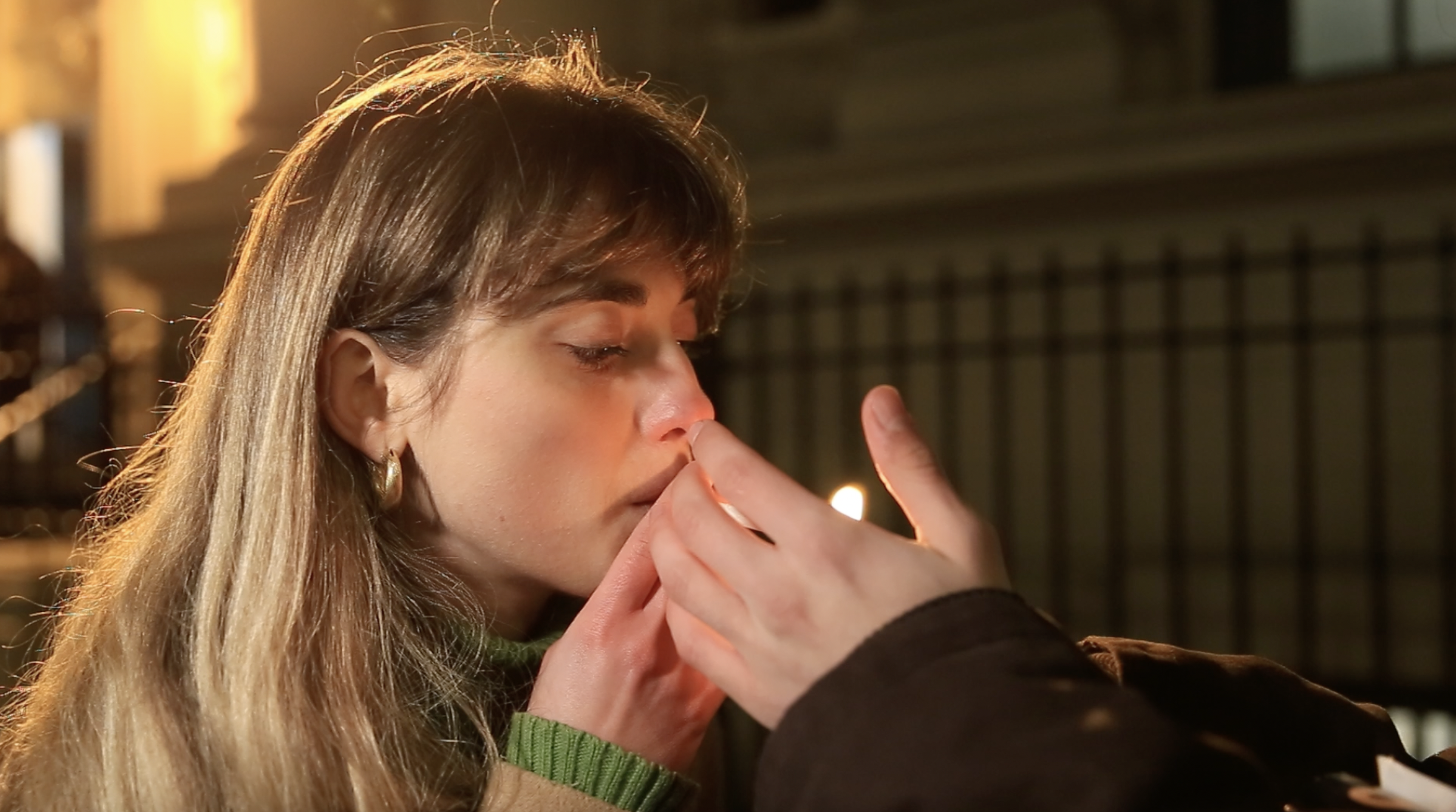 A young woman holds a cigarette up. A man's hands light it for her.