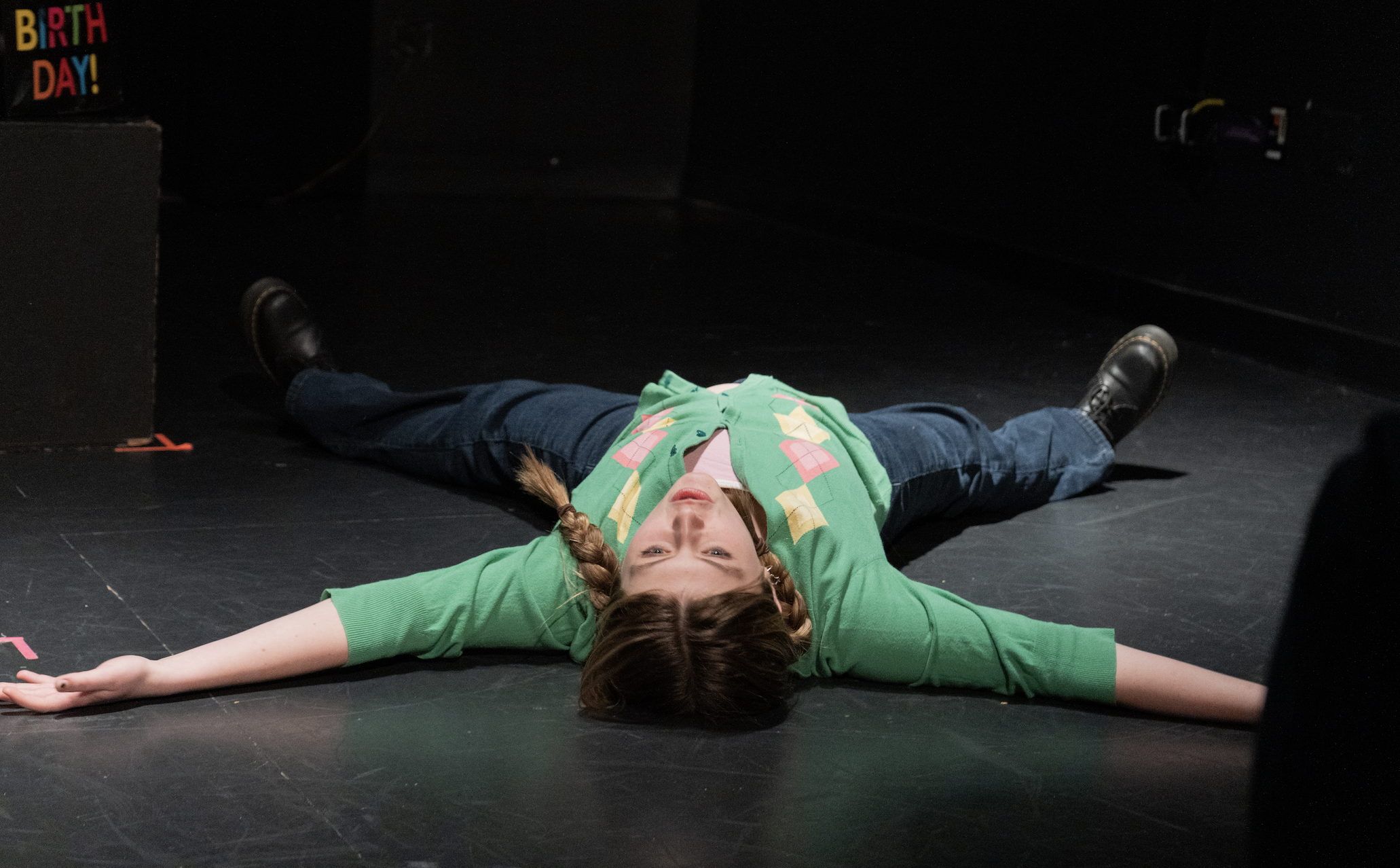 A girl with two braids in her hair lays on the floor of a stage in a starfish position, arms and legs outstretched.