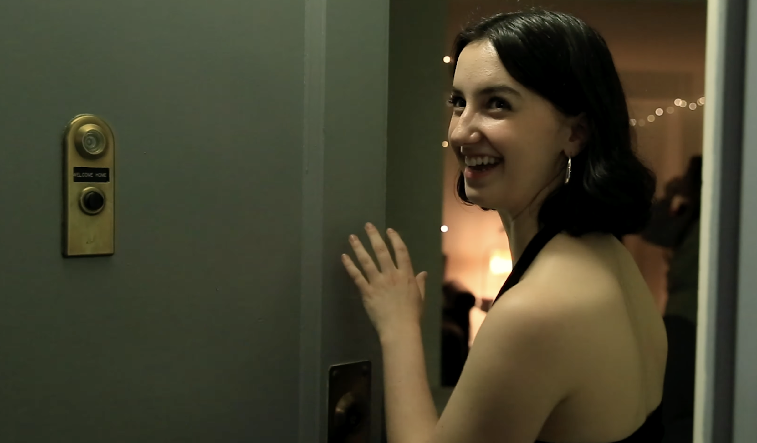 A young woman stands in a door frame, smiling eagerly at someone off-camera. A house party is happening behind her