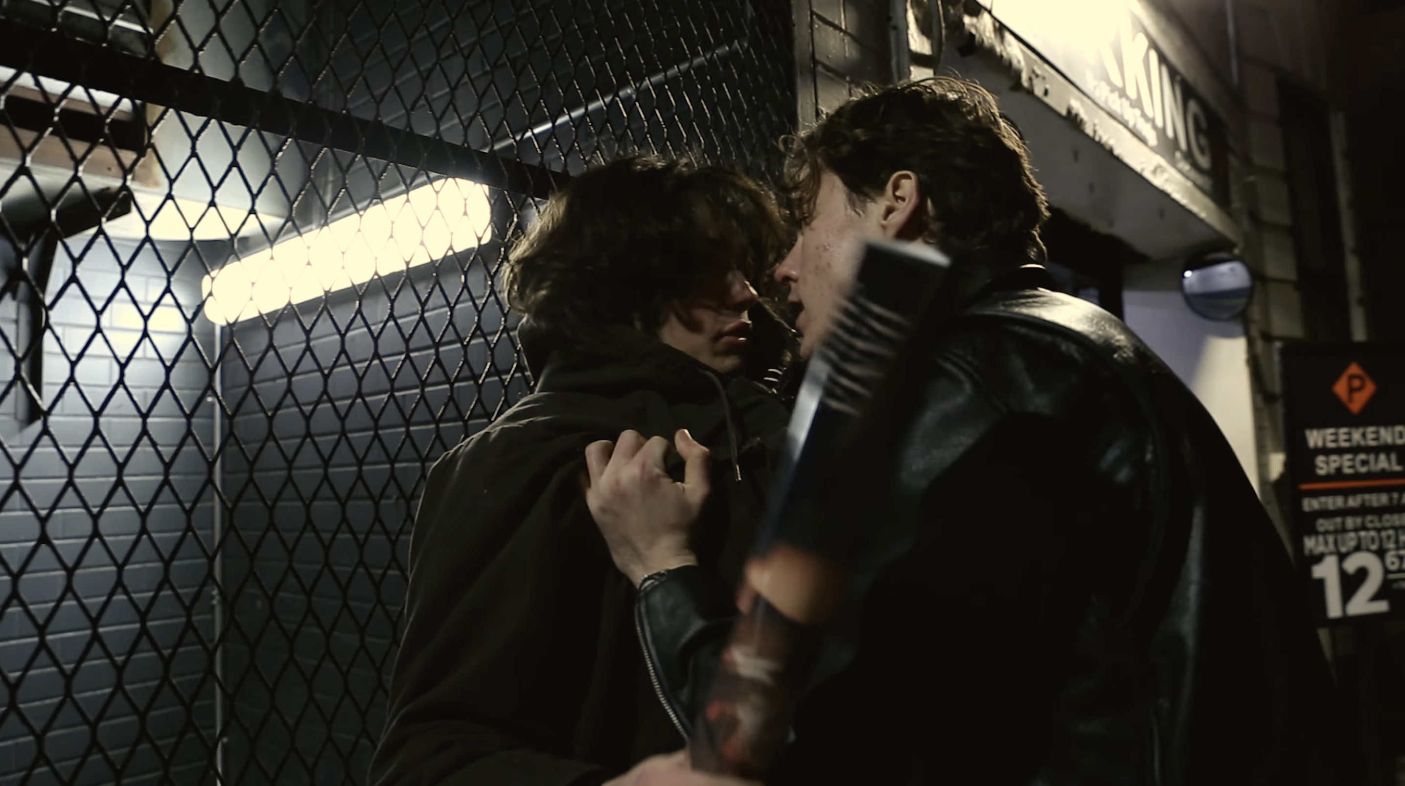 A young man in a leather jacket pushes another against a wire fence in New York City. It is nighttime.