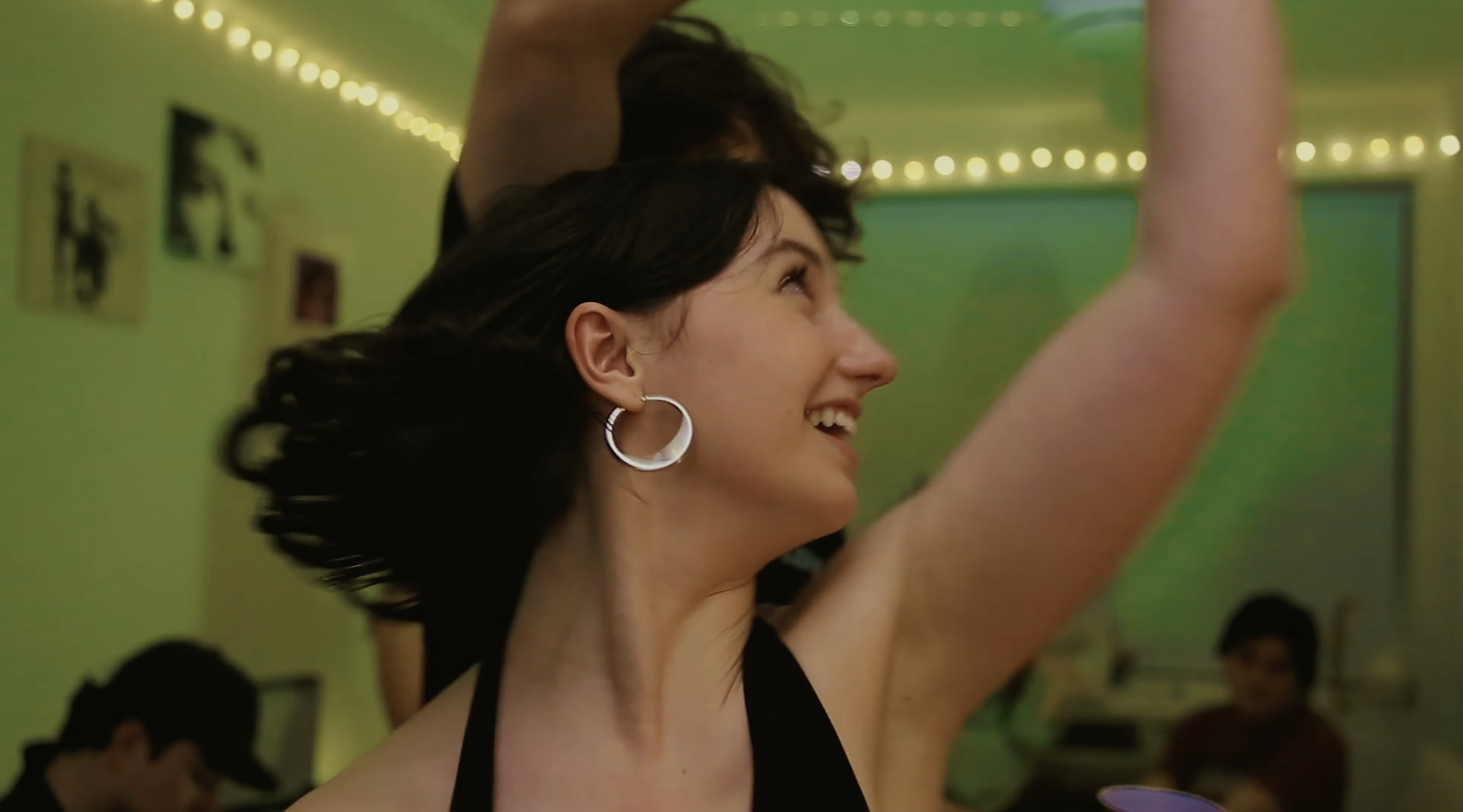 A young woman with short dark hair dances with a young man. They are at house party. The lights are green around them.