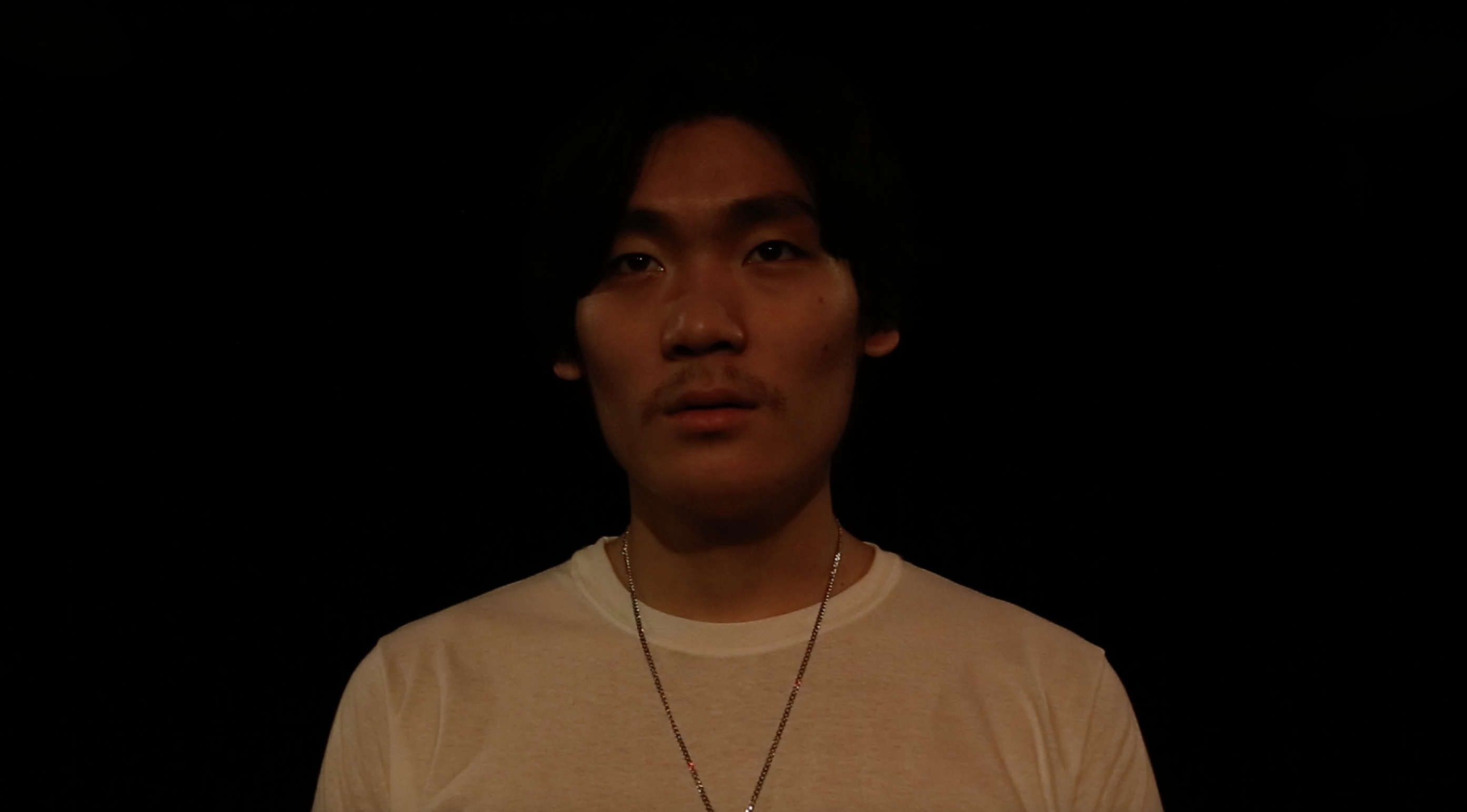 A young man in a white shirt with a silver necklace stares outwards. He has a look of yearning and intrigue