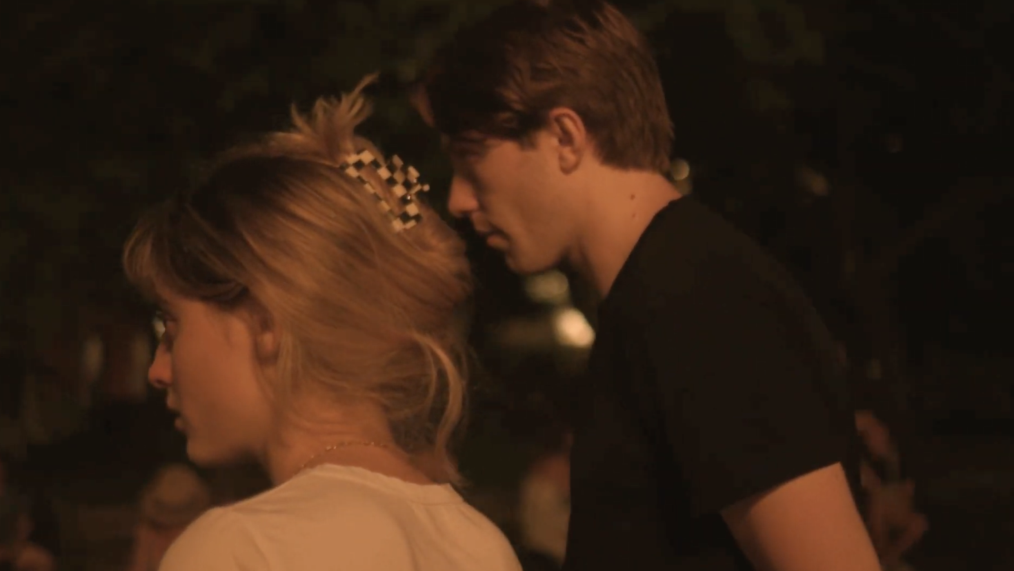 A young man and a young woman stand in a park. Only their side profiles are visible.