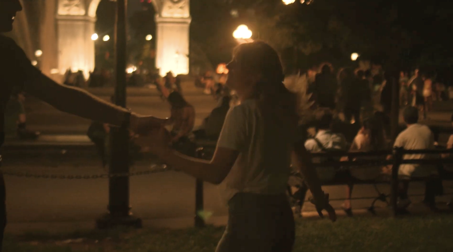 A young woman dances with a young man. The arch in Washington Square Park is shining behind them. The park is full of people.