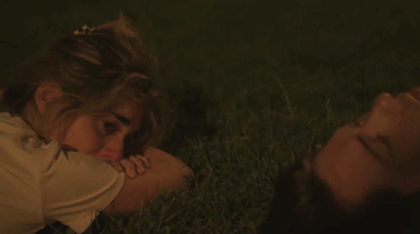 A young woman lies in the grass and cries. A man lies next to her stargazing, and is unaware of her sadness.