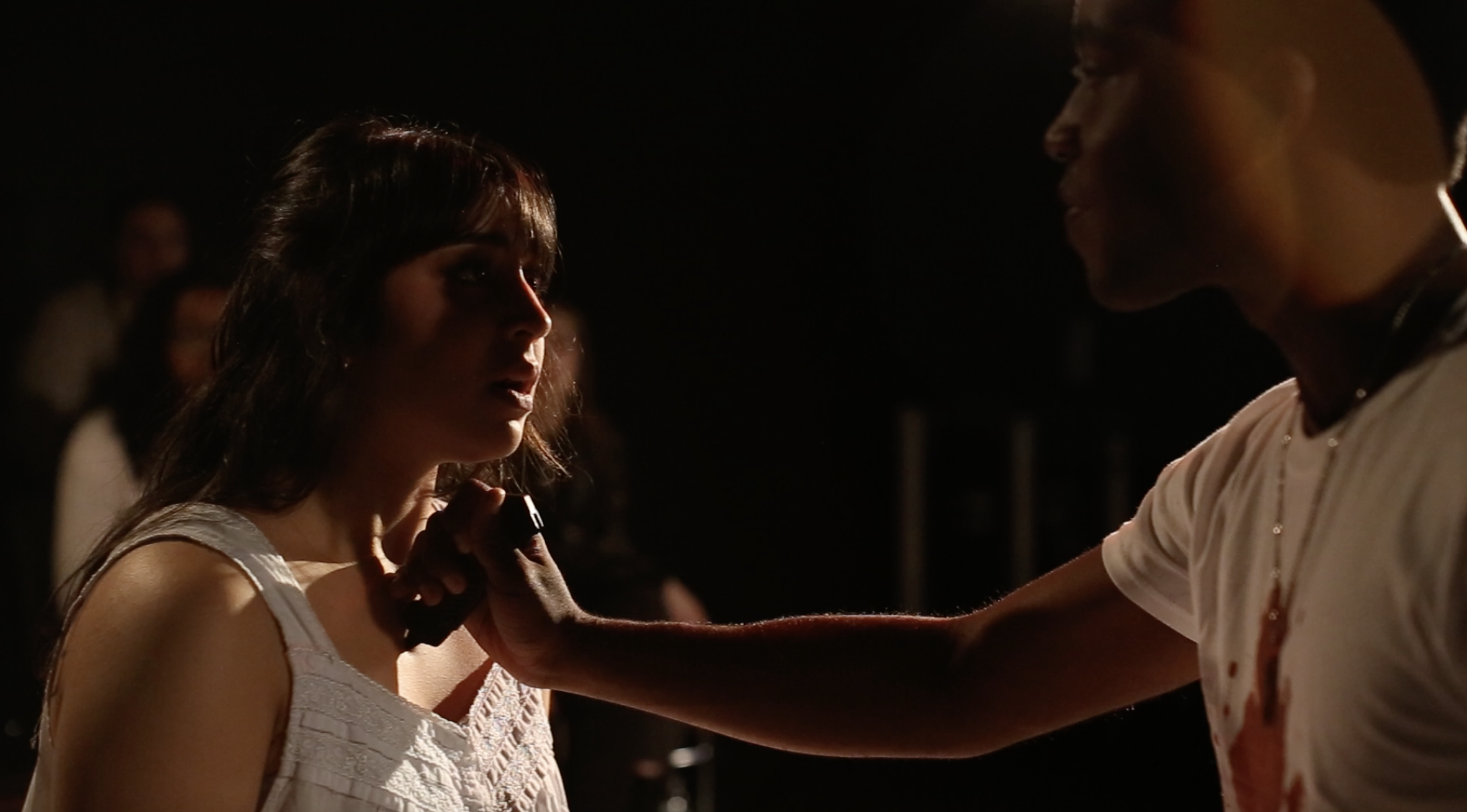 A young actor with a prop knife presses it into a young woman's chest. An audience watches them