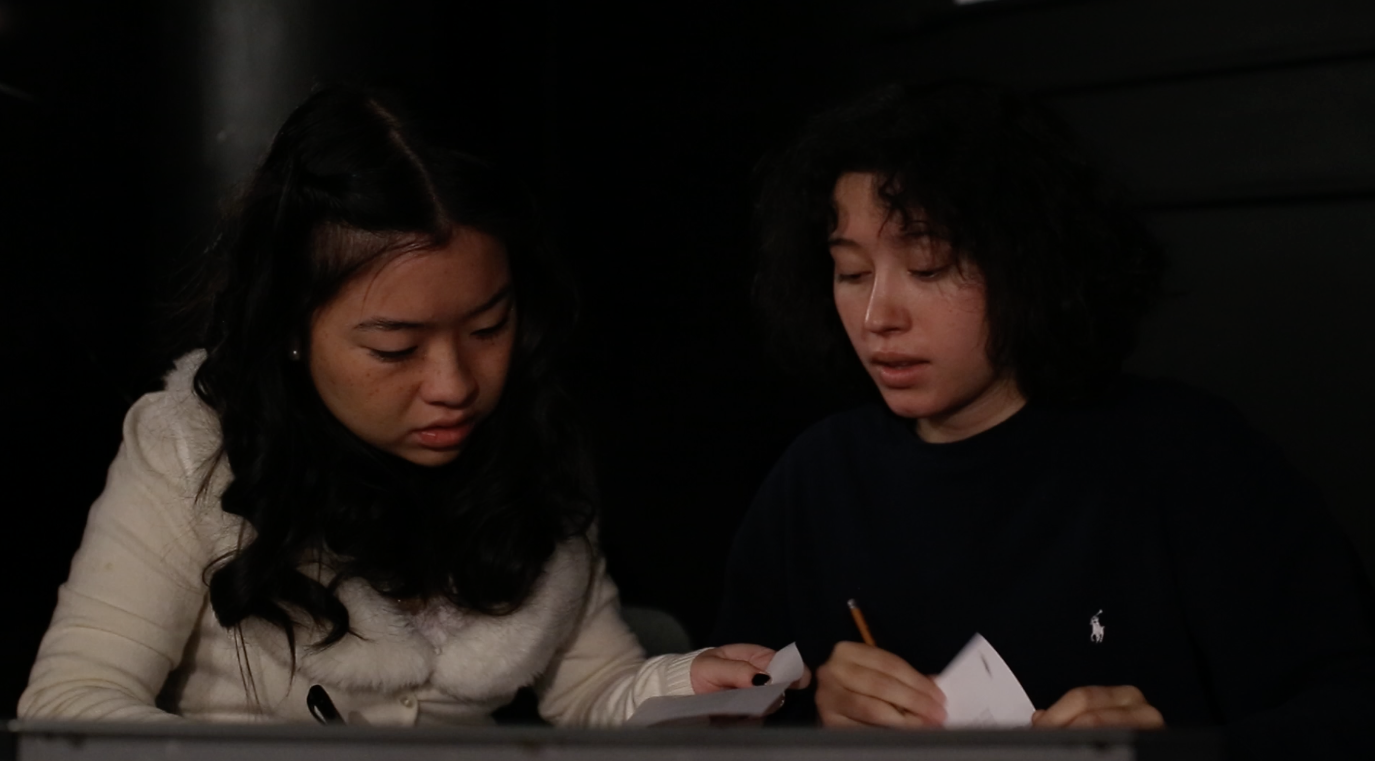 Two young women sit at a lighting booth in a theater. They stare at a script with confusion.