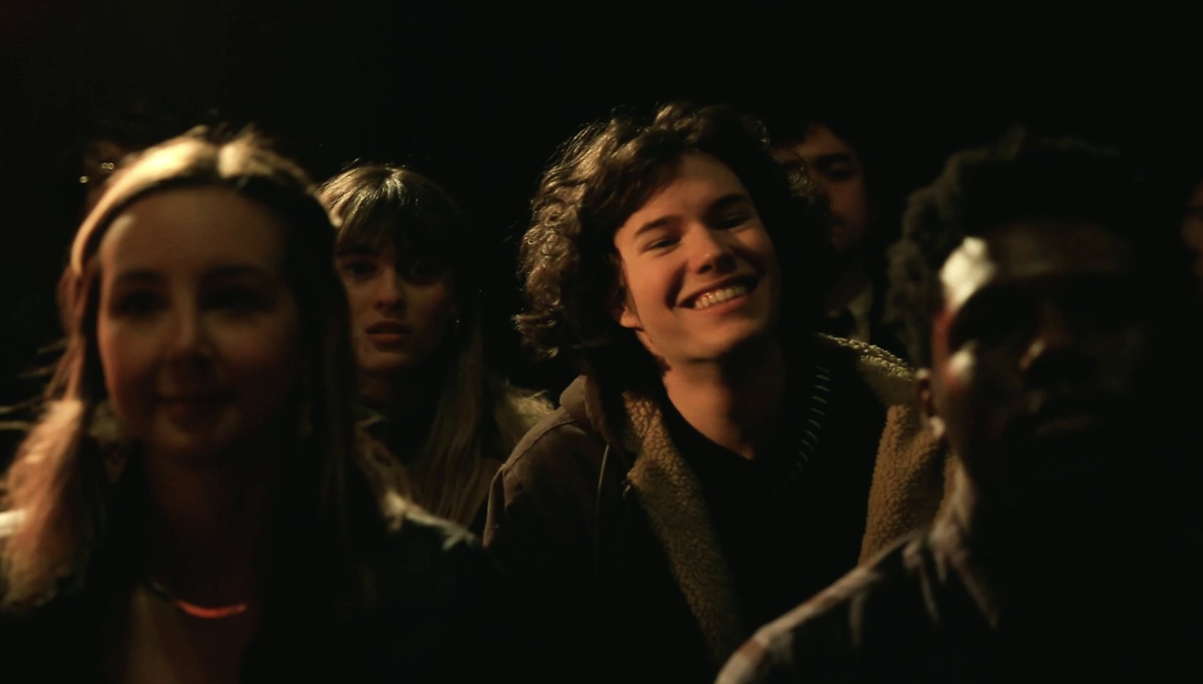 A young man sits in an audience. He beams at someone on-stage that is out of frame.