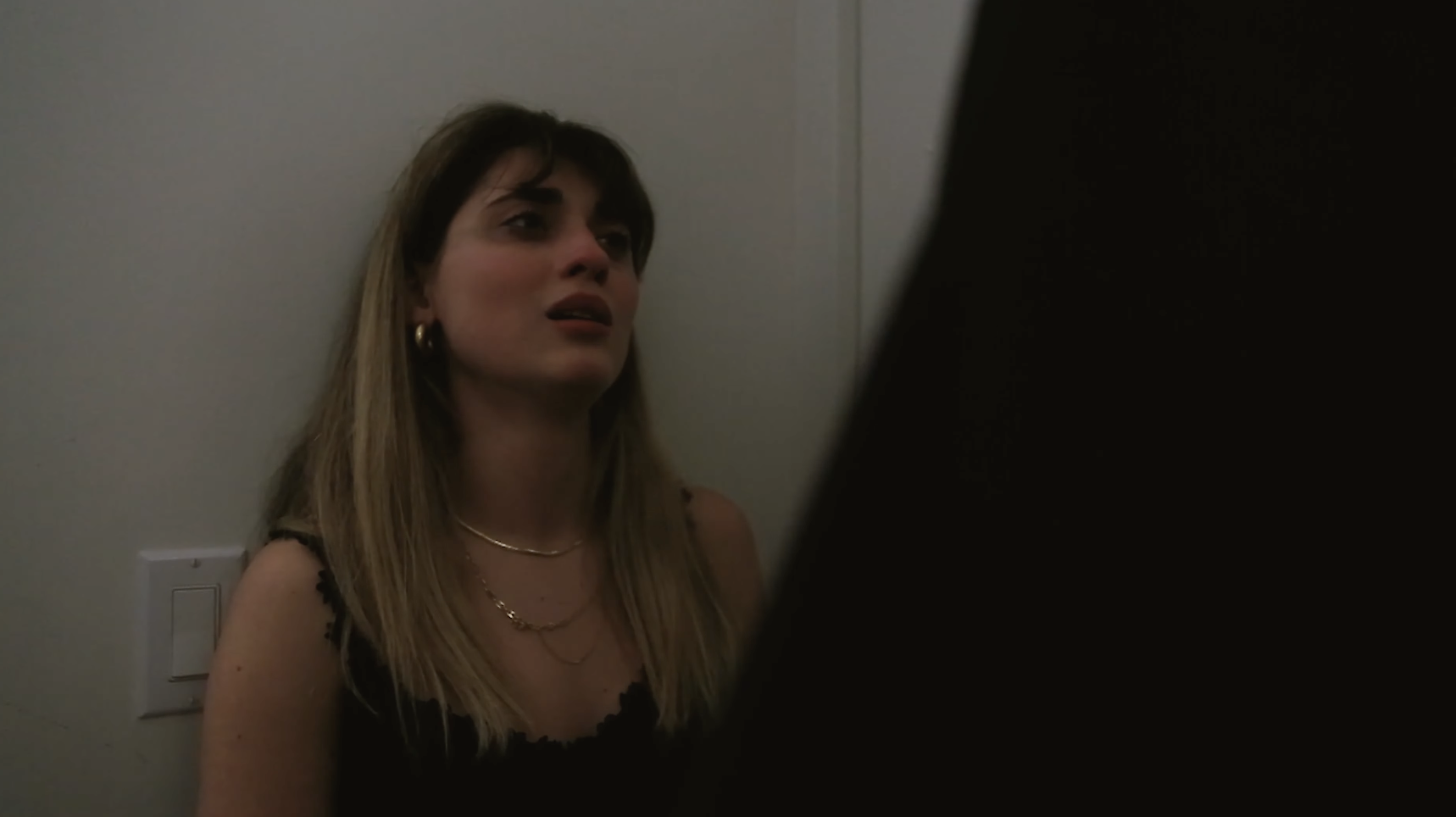 A young woman stands against a wall with a teary look.
