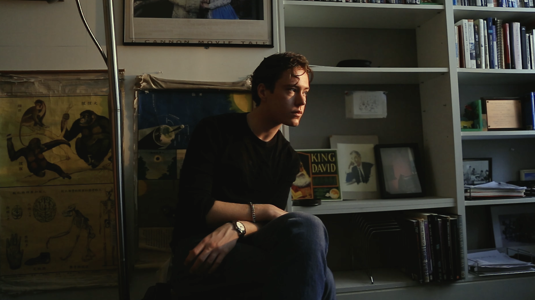 A young man sits in an office, the sun beaming on his face. His legs are crossed.