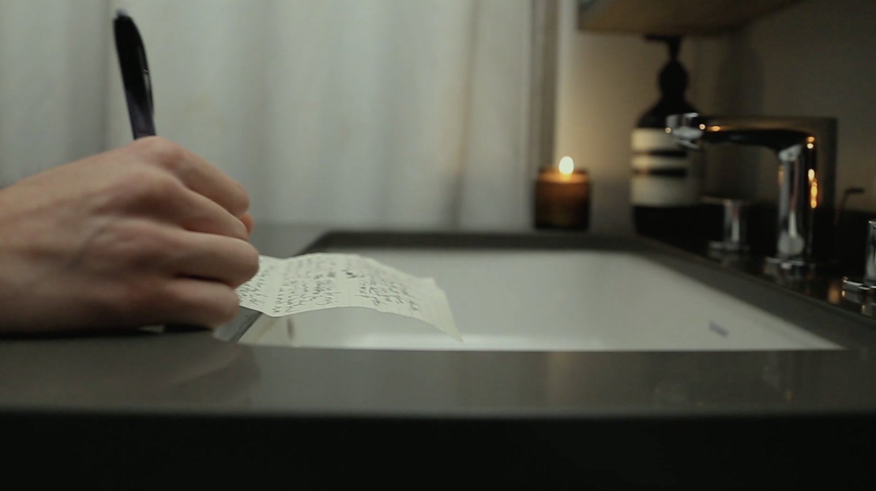 A hand scribbles on a letter. A candle is lit behind it.