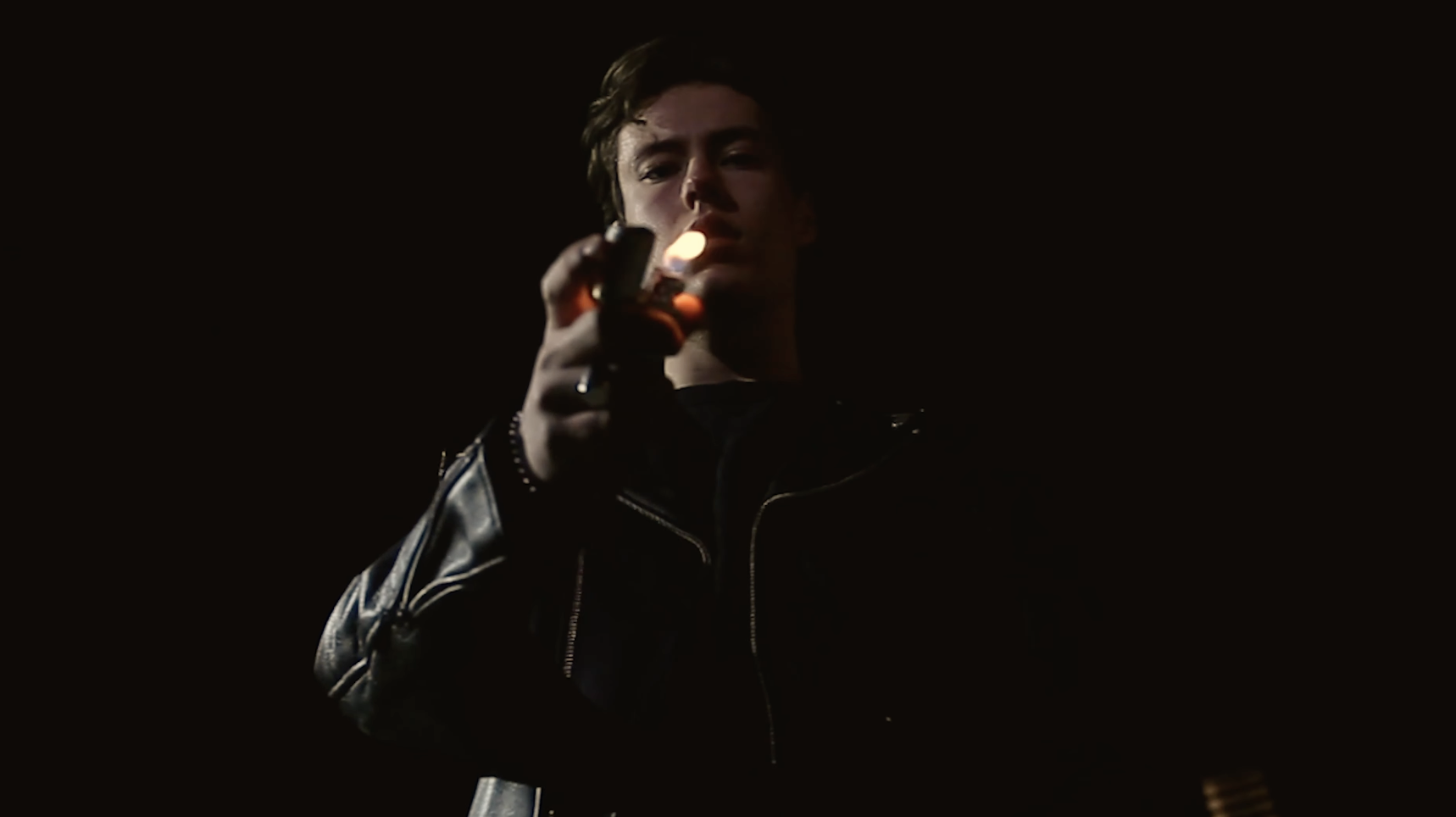 A young man in a leather jacket holds a lighter. He stares down with a mixture of concern and confusion.