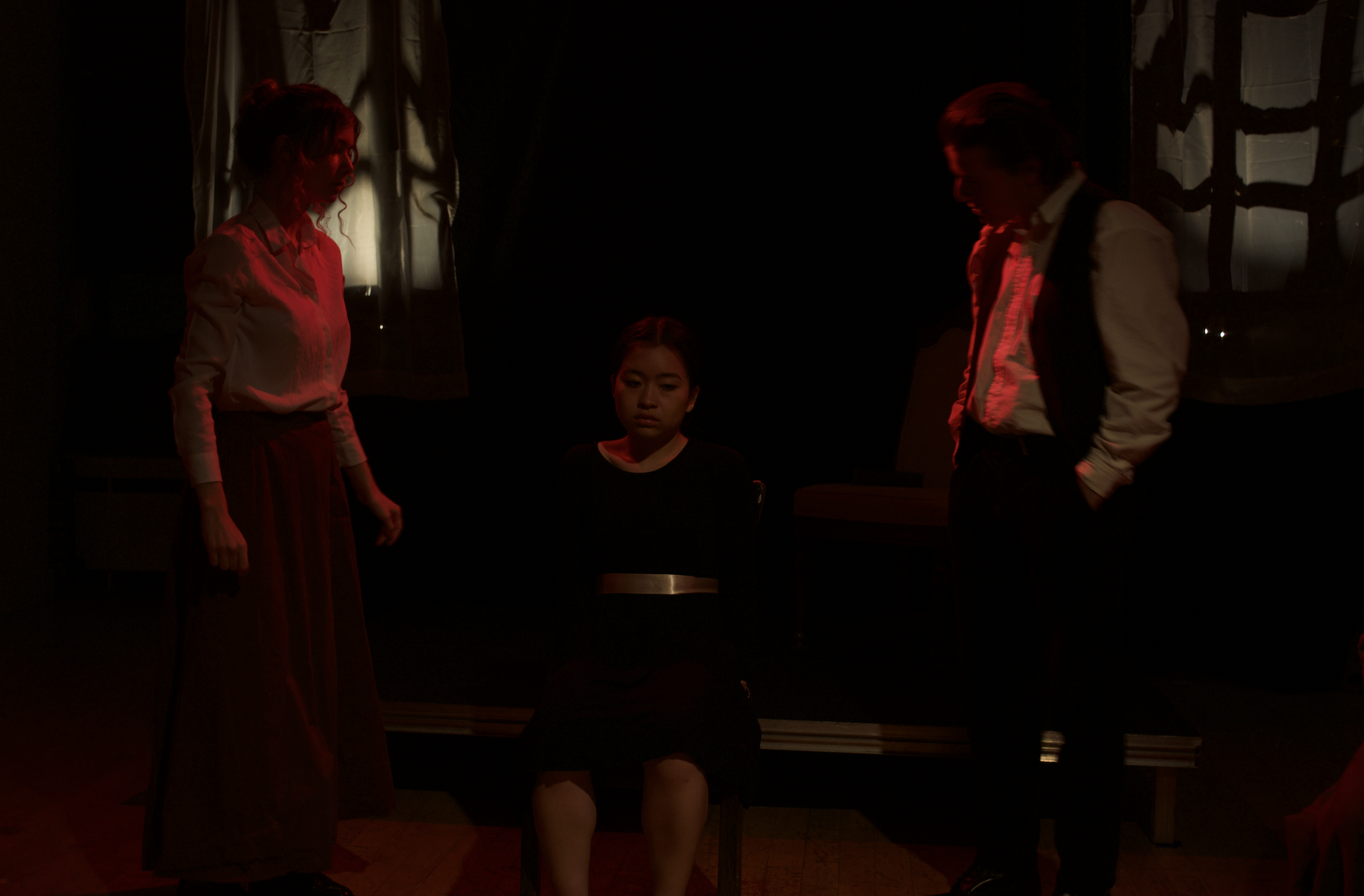 Jane Eyre (2022) A young actress sits in a chair while two older-looking actors stand over her menacingly. The lights are red around them.